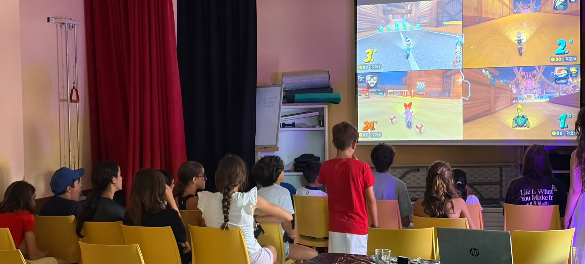 Niñas, niños y jóvenes sentados frente a una pantalla grande con una partida a cuatro jugadores en Calahorra de Boedo